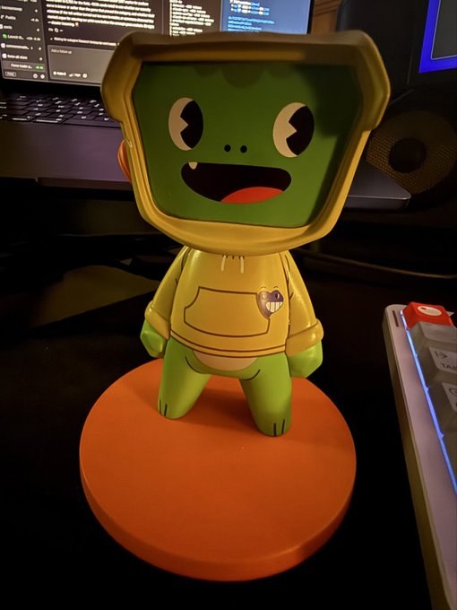 A cute green cartoon character figurine wearing a yellow hoodie sits on an orange circular base on a desk, with monitors and a keyboard visible in the background. The post suggests this NFT-style figurine was gifted from "Blokyz," possibly indicating whitelist eligibility.