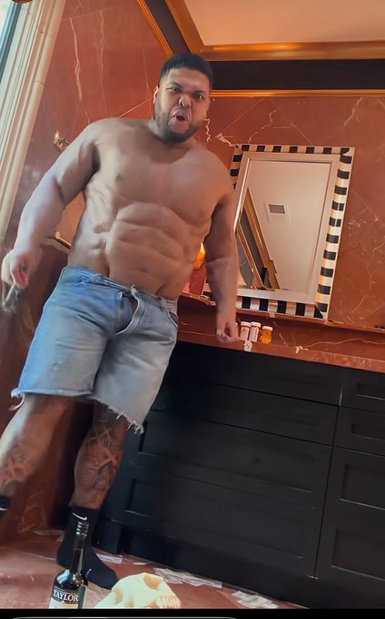 A muscular shirtless man in cutoff denim shorts flexes aggressively toward the camera in a bathroom, with a Bell's whisky bottle and cap on the counter near his foot. The image humorously illustrates the post's "CRO?!?!!!!!!" reaction, using his intense, bulging physique and scowling expression to dramatize shock and frustration.