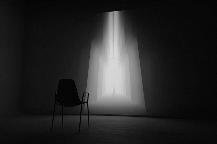 The image features an empty room with a chair positioned near a large window that allows light to shine through. The chair appears to be made of metal or plastic, as indicated by its design. The scene seems to be set up for an auction, possibly featuring art pieces related to patience and the concept of time.