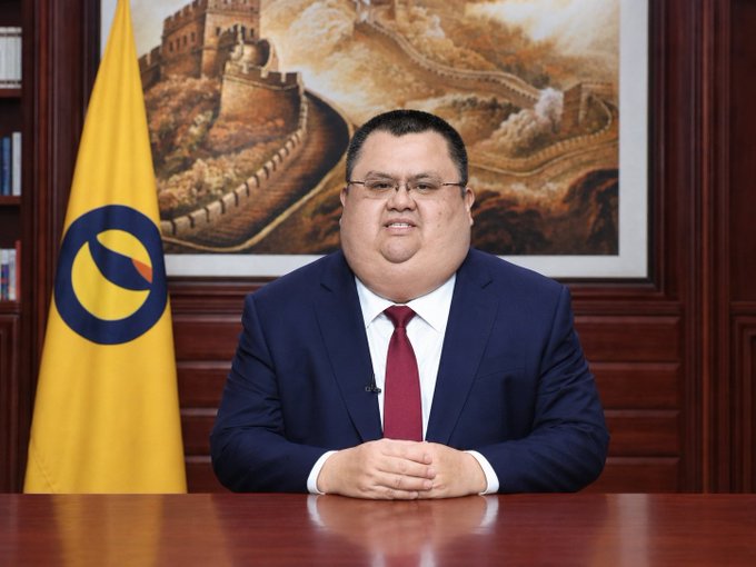A man in a dark suit with a red tie sits at a desk in a formal setting with a yellow flag bearing a circular logo to his left and a large painting of the Great Wall of China behind him.