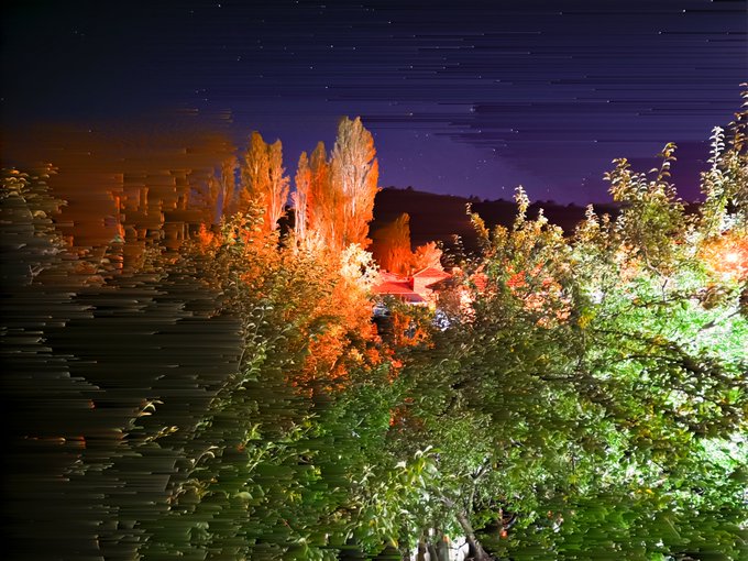 A nighttime photograph with motion blur, showing illuminated trees lit with orange and green lights against a dark sky, with a building barely visible in the background.