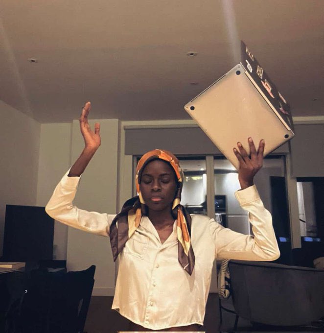 A woman in a silk blouse and headscarf balances a large book labeled "The Bible" on her head while raising her other hand in a prayerful gesture, humorously depicting her solemn appeal to God for help achieving her ambitious September goals of growing her social media following and earning figures from web3, NFTs, and memes.