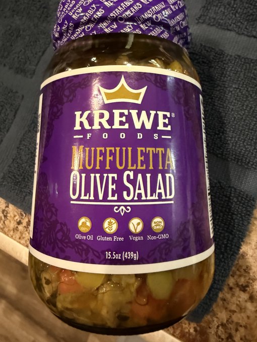 A jar of Krewe Foods Muffuletta Olive Salad (15.5oz/439g) sits on a granite countertop, showcasing its colorful mix of olives and vegetables through the clear glass—perfect for the mouth-watering anticipation of using this olive oil-based, gluten-free, vegan, and non-GMO spread.