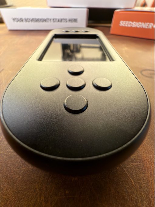 A black handheld electronic device with a screen and directional buttons rests on a wooden surface, with boxes labeled 'YOUR SOVEREIGNTY STARTS HERE' and 'SEEDSIGNER' visible in the background.