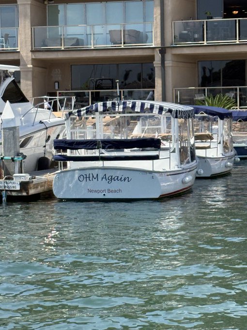 A white boat named 'OHM Again' from Newport Beach docked in a marina, with other boats and a waterfront building visible in the background.