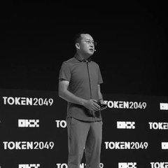A black-and-white photo shows a man presenting on stage at the TOKEN2049 conference, holding a clicker.