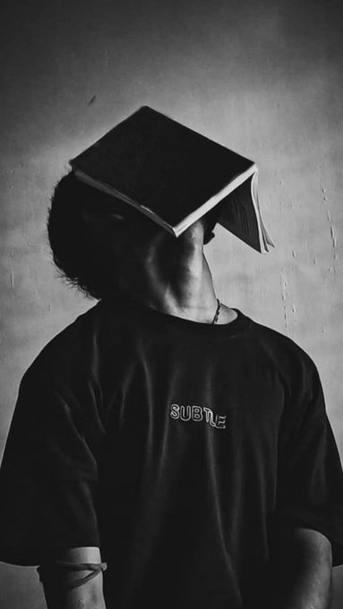 A black-and-white photo of a person wearing a black t-shirt with "SUBTLE" text, their face obscured by an open book balanced on their head.