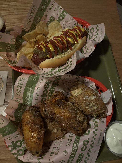 A meal spread featuring a loaded hot dog with ketchup and mustard, potato chips, and seasoned fried chicken wings served in red baskets lined with 'Clean Eatz Kitchen' branded paper.