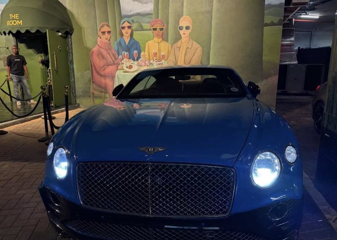 A sleek blue Bentley sports car is parked in front of a vibrant mural featuring four figures in sunglasses, with "THE ROOM" signage visible on a nearby green awning, perfectly embodying the celebratory "Gm Winners" vibe with its luxurious, prize-worthy appearance.
