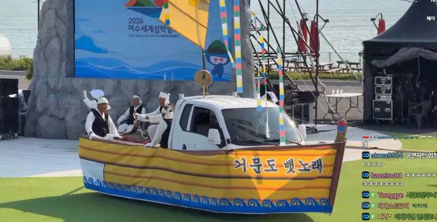 Performers in traditional Korean attire row an oar-less boat mounted on a truck, parading past a "2026 여수세계섬박람회" (2026 Yeosu World Island Expo) banner, while a live chat scrolls reactions on the right.