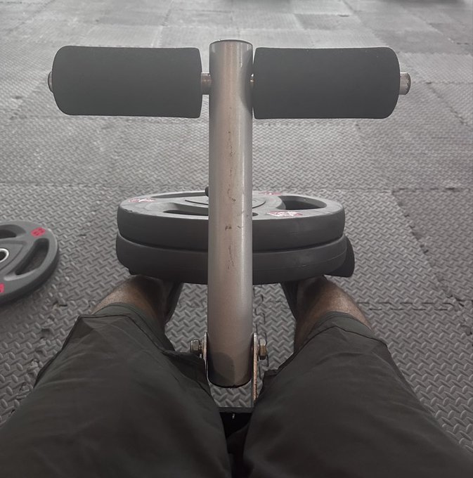 A first-person perspective shows someone sitting on gym equipment with weight plates resting on their legs, looking down at their knees and the padded rollers.