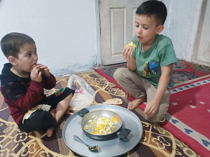 Two young boys sit on a patterned rug, eating eggs from a metal pan placed on a tray between them in a modest home setting.