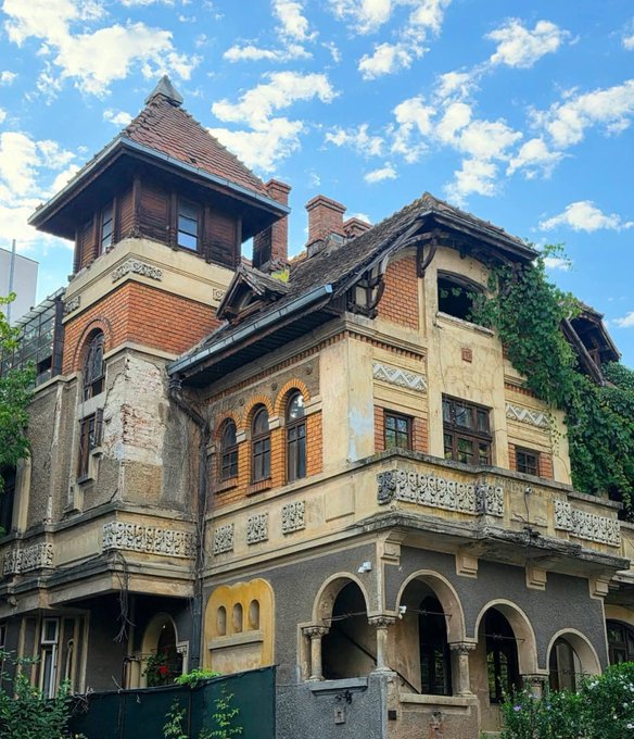 An old, ornate, multi-story brick and stucco building with a tower, arched entryways, and ivy growing on its walls, set against a blue sky with scattered clouds.