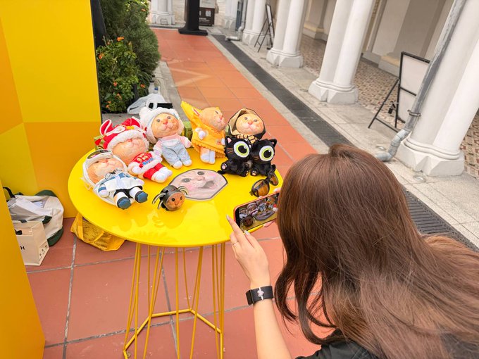 A woman with long brown hair photographs a collection of small plush toys arranged on a bright yellow outdoor table.