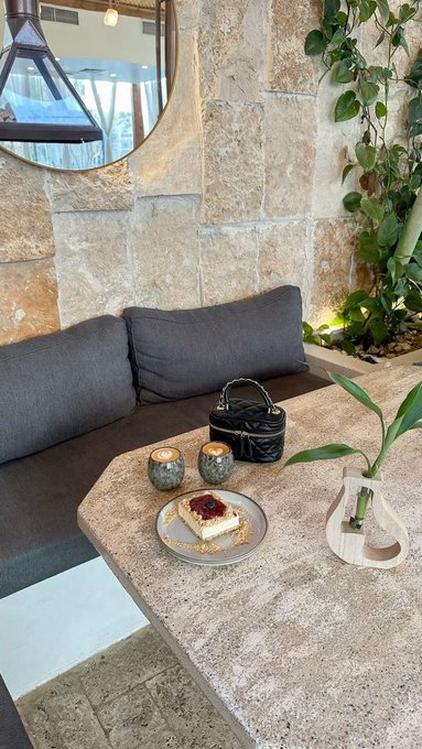 An interior photo of a stylish café or restaurant with a stone wall, a round mirror, a black sofa, and a hexagonal stone table with a slice of cake topped with red jam, two small ceramic cups, and a small black bag, with green plants adding ambiance.