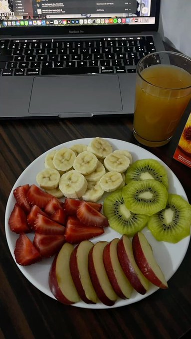 A white plate holds sliced bananas, strawberries, kiwi, and apple arranged in sections, sitting on a dark wooden table next to a MacBook Pro and a glass of orange juice.