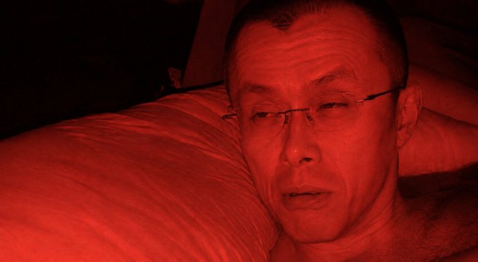A close-up portrait of an older man wearing rimless glasses, lying on a pillow with the entire image bathed in dramatic red lighting.