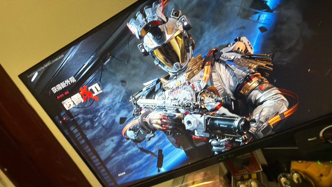 A photograph of a monitor displaying artwork from the video game Apex Legends, featuring an armored character with the Chinese text '坚韧不屈' visible.