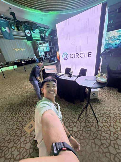 A man takes a selfie at a Circle crypto event booth, with a Hyperliquid banner visible in the background at a conference venue.