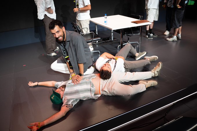Three young men pose playfully on a stage, with one lying on his back while the others lie across him in a star formation, all smiling at the camera.