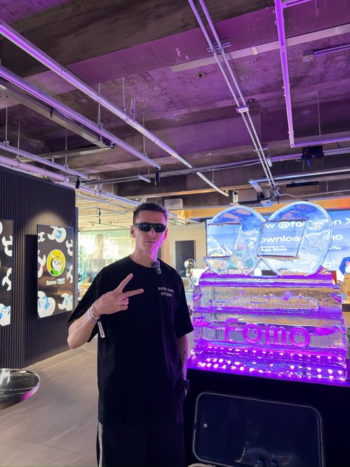 A man wearing sunglasses and a black t-shirt with "WE ARE ETERNITY" poses with a peace sign in a neon-lit indoor venue, standing next to a glowing purple display labeled "KLON".