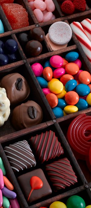 A close-up, vertical view of a wooden box filled with various colorful chocolates and candies, including sprinkled treats, colorful round candies, coffee beans, and drizzled chocolate squares.
