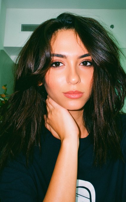 A young woman with long dark wavy hair poses with her hand resting on her chin, wearing a black t-shirt with a partial white logo visible. The image complements the post's playful request for viewers to share their Solana (SOL) crypto wallet addresses in a flirty, casual tone.