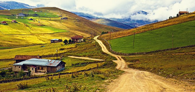 A scenic landscape of rolling green and yellow hills with small rural houses, a winding dirt road, and cloudy skies in the mountains.