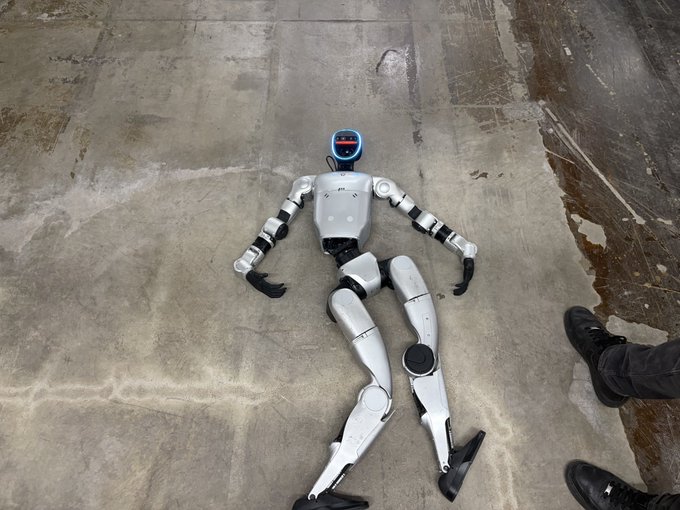 A humanoid robot with a digital display for a face lies sprawled on its back on a concrete floor, next to a person's feet—humorously reinforcing the "AH FUCK" caption by suggesting the bot has just taken a dramatic tumble or malfunctioned.