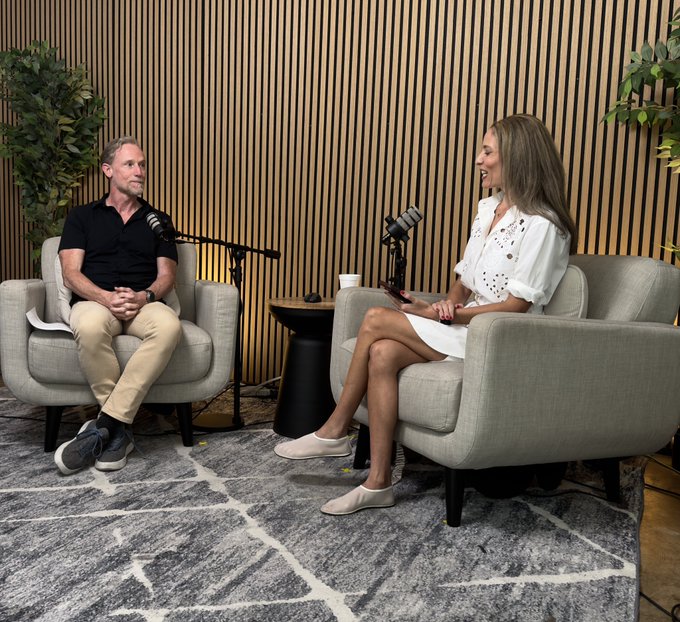 Two people have a conversation in a podcast studio setting with microphones, seated in gray armchairs against a wooden slat wall backdrop.