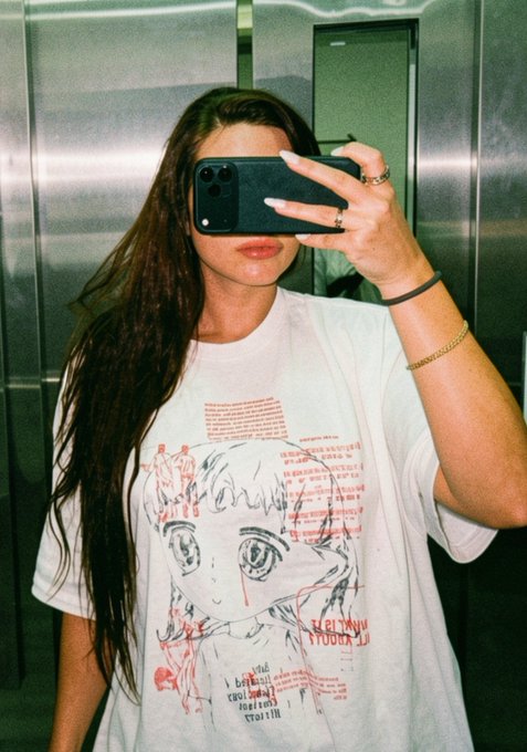 A young woman with long dark hair takes a mirror selfie in an elevator, holding up a black iPhone that obscures her face. She wears an oversized white graphic t-shirt featuring an anime-style character illustration with red text overlays and the word "MILADY" referenced in the accompanying post.