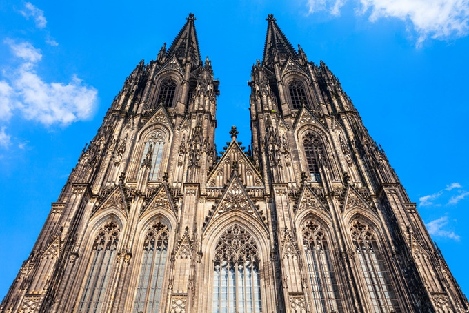 Low-angle photograph of the Cologne Cathedral's Gothic twin spires reaching into a blue sky with scattered clouds.