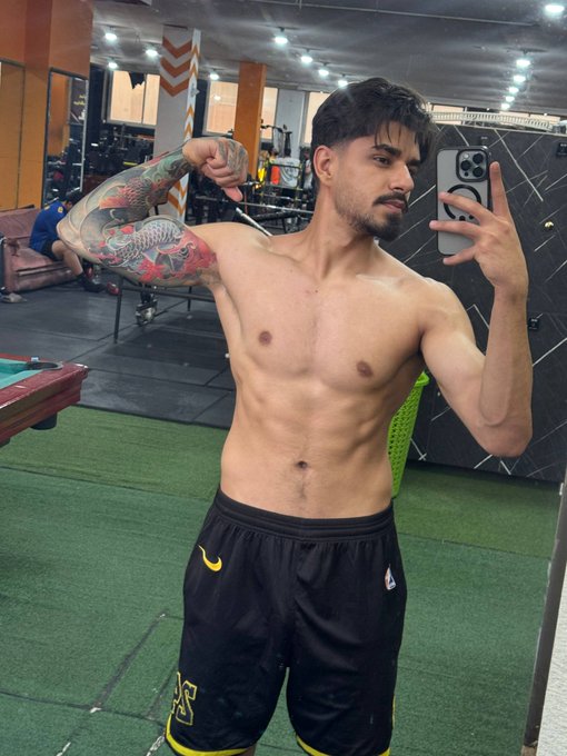 A shirtless young man with tattoos flexes his bicep while taking a mirror selfie in a gym, wearing black shorts with a yellow logo.