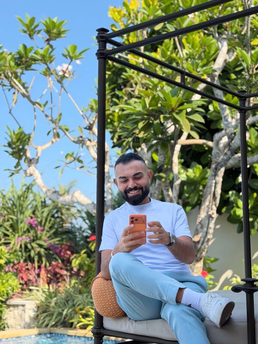 A smiling bearded man sitting on a canopy daybed outdoors in a tropical setting with lush greenery, holding an orange smartphone.