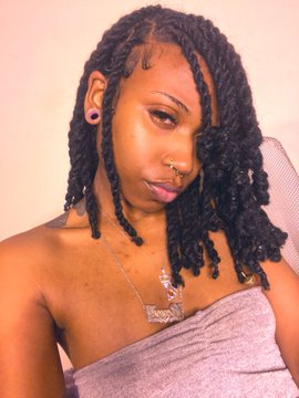 A young woman with shoulder-length twist braids poses for a selfie, wearing a strapless top and a pendant necklace, with a soft, contemplative expression against a plain background. The "꩜" symbol, a Sundanese script character, adds an artistic, enigmatic flair to the minimalist portrait.