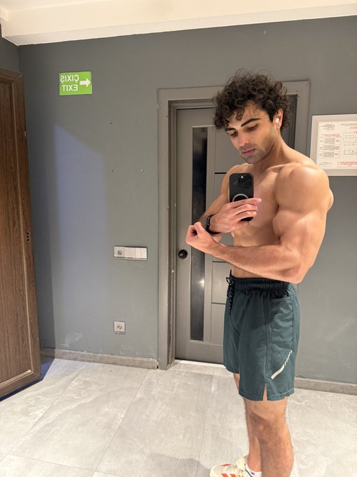 A shirtless man with curly hair takes a mirror selfie while flexing his arm, standing in a hallway near an exit door.