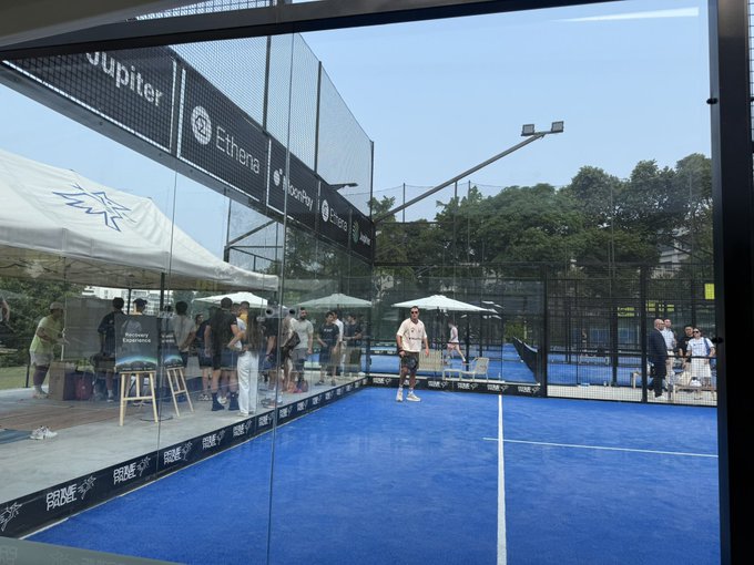 A padel match is underway at the Ethena Padel court, with players on the blue court and spectators gathered under white tents nearby. Sponsor banners reading "Jupiter," "Ethena," and "BlackBerry" line the glass walls, fitting the post's mention of being at Ethena Padel.