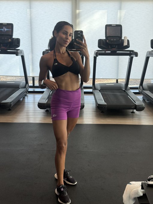 A woman taking a mirror selfie in a gym, wearing a black sports bra and pink shorts, with treadmills visible in the background.