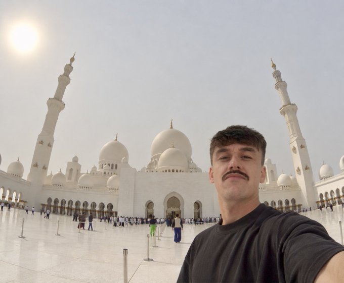 A selfie of a young man with a mustache in front of the Sheikh Zayed Grand Mosque, with the sun shining brightly in the sky.