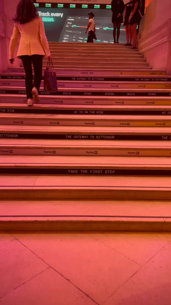 The image shows a group of people walking up a set of stairs with a large screen displaying information about the opening ceremony at the Louvre Museum. The stairs are adorned with black and white stripes, adding an artistic touch to the scene. A woman is seen carrying a handbag as she ascends the stairs, while another person can be spotted in the background. The image also includes text that reads "TAO - @const_reborn & @markjeffrey opening @proofoftalk main stage at the Louvre, for the second and last fay of the festival. And yes, Bittensor is everywhere!".