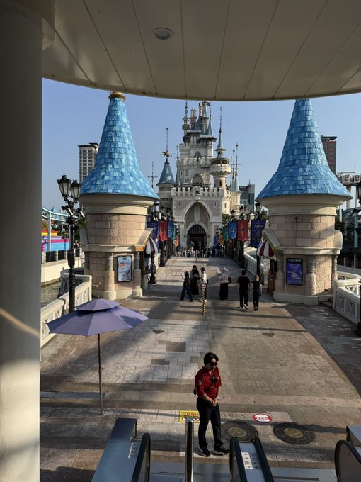 A view of Lotte World castle entrance in Seoul, featuring blue-turreted towers and a security guard wearing a mask standing at the bottom near escalators.