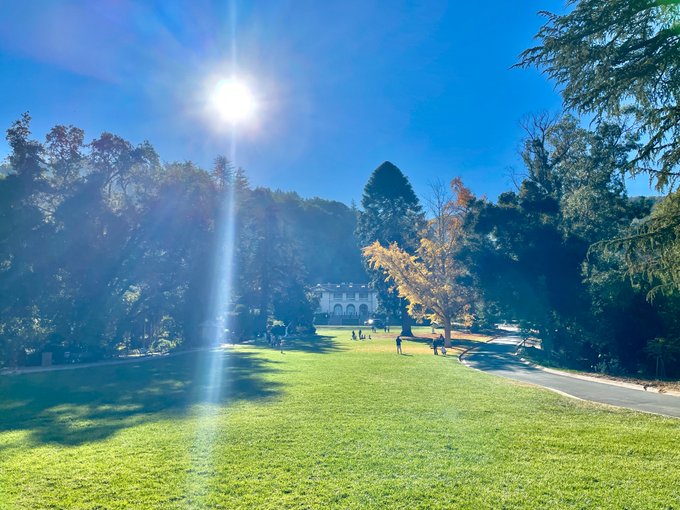 A scenic landscape features a large grassy field bathed in sunlight, with people walking toward a white building framed by trees under a bright blue sky.