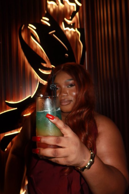 A young woman with long reddish-brown hair and bold makeup poses with a green cocktail, showing off her bright red manicure and gold bracelet against a dramatic background with illuminated flame-like designs. The stylish, confident pose with her cocktail in hand complements the post's playful Pidgin English caption about shining her teeth in a vibrant nightlife or lounge setting.