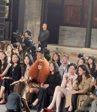 An audience seated at a fashion show or event, with a woman in the front row wearing a large, voluminous red fur stole, while a man in a suit stands in the background near a camera setup.