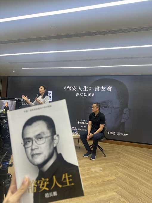 A book signing event for 《幣安人生》 (Binance Life) where the author 趙長鵬 (CZ) sits on stage while someone holds up a copy of the book featuring his portrait on the cover.