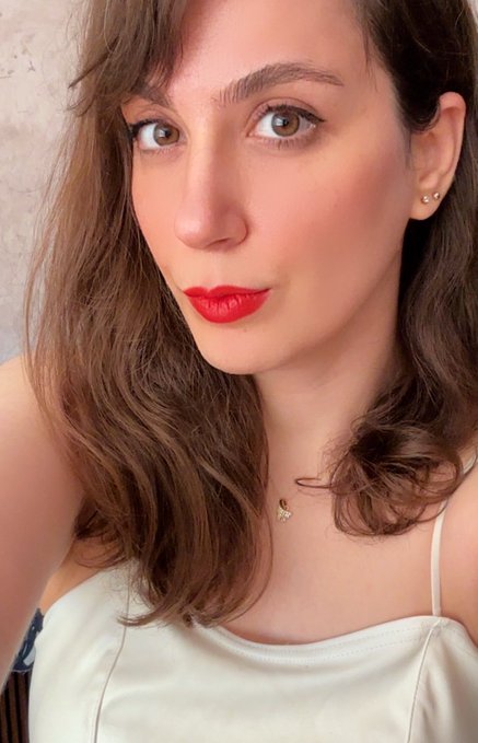 A close-up selfie of a woman with brown wavy hair and bangs, wearing bold red lipstick, a white top, and a small gold pendant necklace.