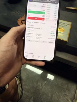A person holds a smartphone displaying a trading or portfolio app showing detailed account information with green and red figures, likely reflecting recent stock gains and losses. The phone screen captures the dread of checking their holdings during a volatile period with the stock down about 1.9%.