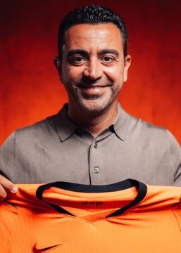 A man in a grey polo shirt smiles while holding up an orange Nike sports jersey against a red background.