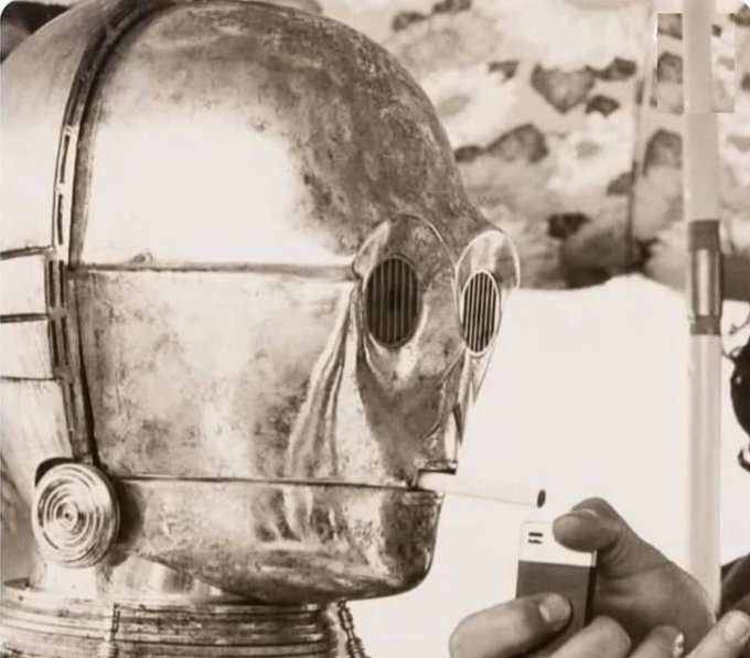 A sepia-toned image of a robot in a metallic helmet-like head holding what appears to be a cigarette or small object.