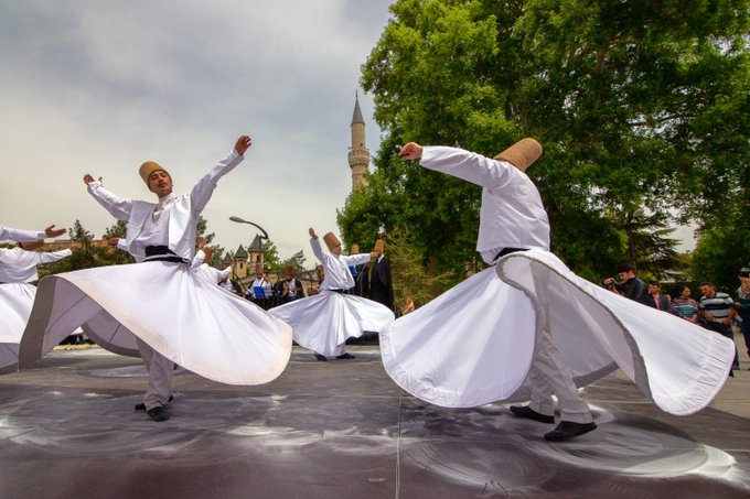 Whirling dervishes in white robes and tall hats perform a traditional Sufi dance outdoors, with one dancer in the foreground on a stage and a minaret visible in the background.